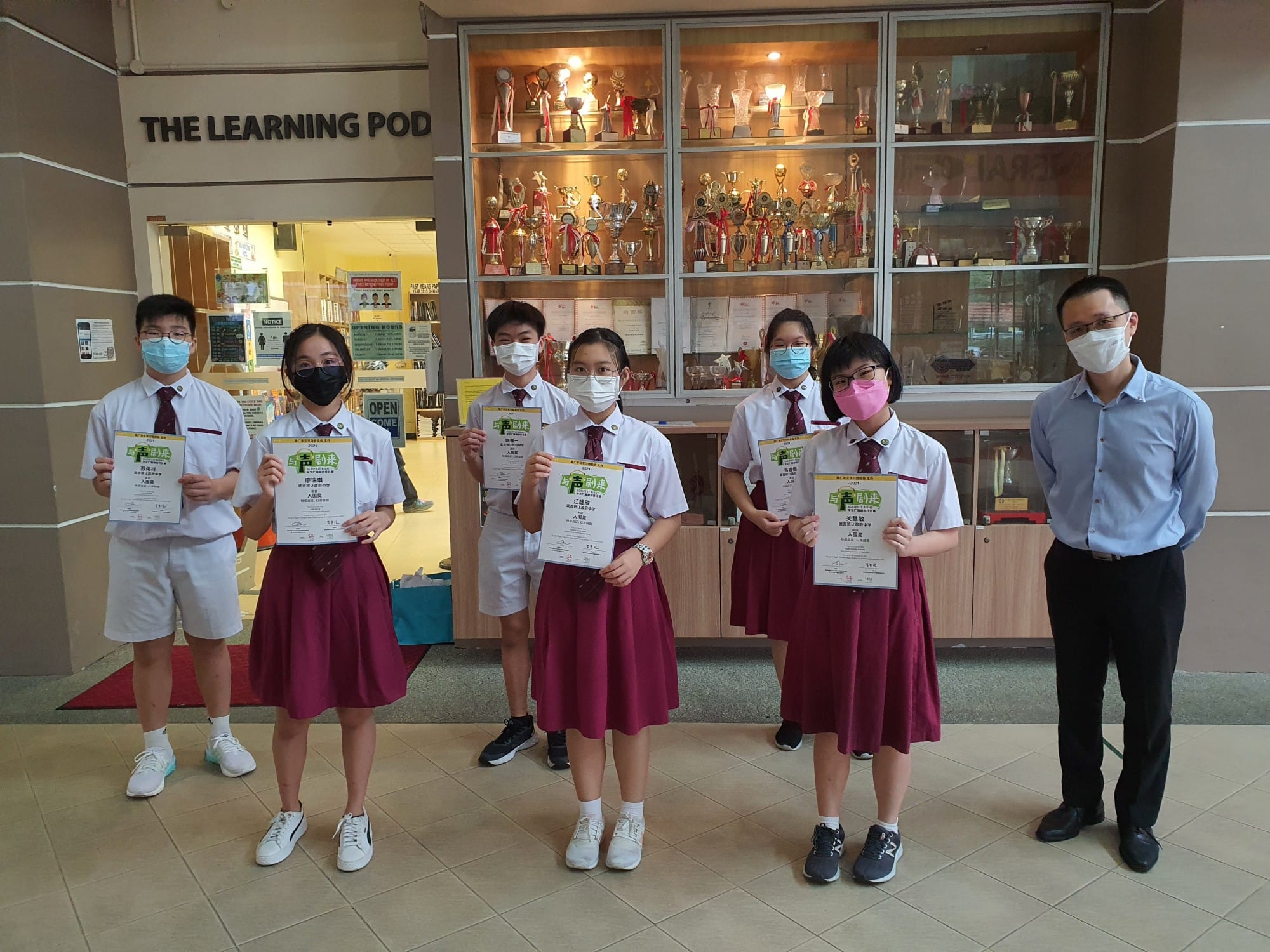 Five masked students in school uniforms hold certificates beside a teacher in front of a trophy display case.